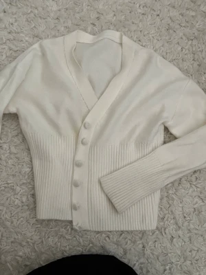 Vit ribbad kofta med knappar - Stilren vit kofta med ribbade detaljer nedtill och vid ärmsluten. Koftan har v-ringning och stängs med matchande vita knappar framtill. Perfekt för lager på lager och enkel att styla till olika outfits.