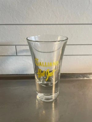 Galliano Hot Shot glas - Säljer ett Galliano Hot Shot glas med gul text. Glaset är transparent och har en klassisk shotglasform som smalnar av mot botten. Perfekt för att servera Hot Shots eller andra drinkar.