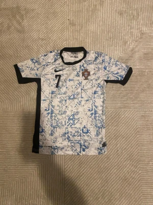 Portugal Ronaldo matchtröja Nike - Säljer en Portugal matchtröja från Nike med nummer 7 och Ronaldo på ryggen. Tröjan är vit med blått mönster, svarta detaljer och korta ärmar. Perfekt för fotbollsfans som vill sticka ut på planen eller läktaren.