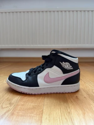 Nike Air Jordan 1 Mid svart/vit/rosa - Snygga Nike Air Jordan 1 Mid sneakers i svart och vitt läder med rosa swoosh. Klassisk siluett med högt skaft och ikonisk Air Jordan-logga på sidan. Perfekta för dig som vill sticka ut med stilrena färgblock och cool design Insidan av sulan har blivit något avskavd (se bild) dock inget som syns vid användning. Denna modell är svår att få tag i idag.
