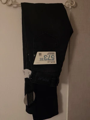 Svarta Replay 573 Bio jeans - Snygga svarta jeans från Replay, modell 573 Bio. Jeansen är tillverkade i ekologiskt material och har klassisk femficksdesign. Märket Replay syns på bakfickan och etiketten. Perfekta för dig som gillar stilrena och hållbara jeans.