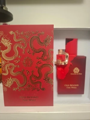 Oud Beyond The Wall - Lyxig parfym från Ministry of Oud i en röd, fyrkantig flaska med gulddetaljer och drakmotiv. Flaskan och förpackningen har eleganta guldmönster och text. Perfekt för dig som gillar exklusiva och orientaliska dofter.