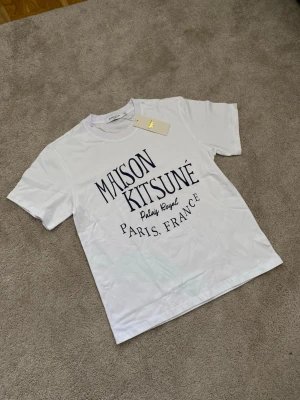 Maison Kitsuné vit t-shirt med tryck - Vit t-shirt från Maison Kitsuné med stort svart tryck på framsidan där det står 'Maison Kitsuné Palais Royal Paris, France'. Klassisk rund hals och korta ärmar. Perfekt för dig som gillar stilrena och trendiga plagg med fransk vibe.