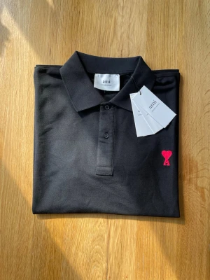 Svart polotopp från Ami med hjärta - Svart polotopp från Ami med klassisk krage och knappar framtill. På bröstet finns ett broderat rött hjärta med bokstaven A. Materialet är mjukt och känns som bomull, perfekt för en clean och stilren look.