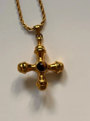 Guldigt halsband med kors och pärla från Prydligt.se - Snyggt halsband med ett korsformat hänge i guldfärgad metall och en mörk pärla i mitten. Kedjan är tvinnad och har ett klassiskt lås. Perfekt för dig som gillar unika accessoarer med lite vintagekänsla.