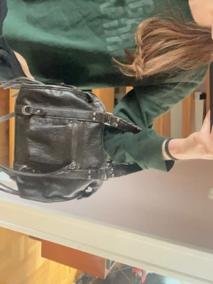 svart handväska med detaljer  - Cool svart handväska i skinnimitation med flera dragkedjor och detaljerade remmar. Väskan har justerbar axelrem och dekorativa metalldelar. Perfekt för dig som gillar en edgy och stilren look. Då väskan är menad att se vintage ut så är den nästan lite brun, men står svart på hemsidan. 💗