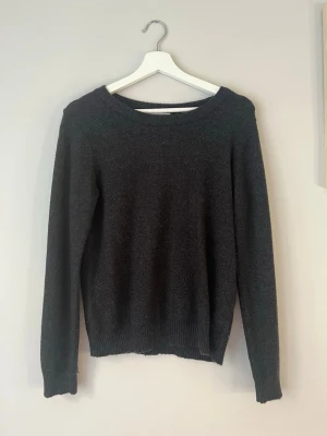 Mörkgrå stickad tröja - En enkel och stilren mörkgrå stickad tröja med rund halsringning och långa ärmar. Tröjan har ribbade muddar vid ärmslut och nederkant. Perfekt att matcha med jeans eller kjol för en avslappnad look.