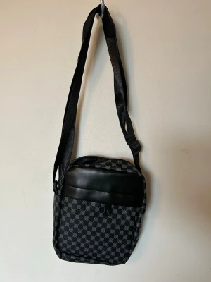 Svart axelväska med rutmönster - Snygg svart axelväska med justerbar rem och rutmönstrad design. Väskan har en framsida i svart konstläder och ett stort huvudfack med dragkedja. Perfekt för att bära det viktigaste och passar till de flesta outfits.