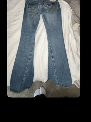 Blå bootcut jeans från True Religion - Säljer ett par ljusblå bootcut jeans från True Religion med klassiska kontrastsömmar och detaljerade fickor bak. Jeansen har låg midja och är tillverkade i denim med stretch för skön passform. Perfekta för dig som gillar retrovibbar och markerade fickor.