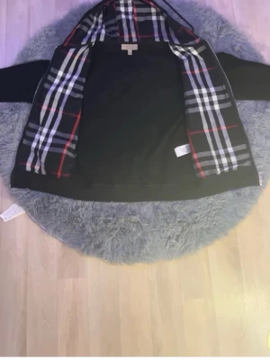 Svart hoodie med dragkedja Burberry - Svart hoodie från Burberry med dragkedja och fickor framtill. Insidan av huvan har det klassiska Burberry-rutiga mönstret i svart, vitt och rött. Justerbar huva med snören och ribbade muddar. Perfekt för en stilren och avslappnad look.