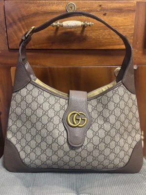 canvas handväska brun - Snygg handväska klassiskt GG canvasmönster med bruna läderdetaljer och guldfärgad GG-logga framtill. Väskan har en axelrem och stängs med ett lock. Perfekt accessoar för dig som gillar lyxig och tidlös design.