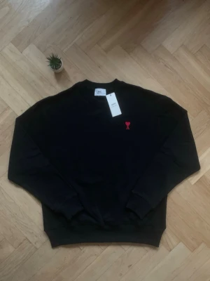 Svart sweatshirt från Ami Paris - Svart sweatshirt från Ami Paris med klassisk rund halsringning och ribbade muddar. På bröstet finns det ikoniska röda Ami de Coeur-broderiet. Tröjan är tillverkad i mjuk bomull och har en avslappnad passform.