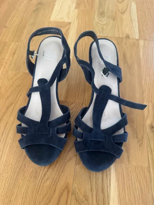 Mörkblåa mocka-pumps från Diasa - Snygga mörkblåa pumps från Diasa i mjuk mocka med öppen tå och remmar över foten. Skorna har en medelhög klack och justerbart spänne vid ankeln. Perfekta för dig som gillar stilrena och trendiga skor med en twist.