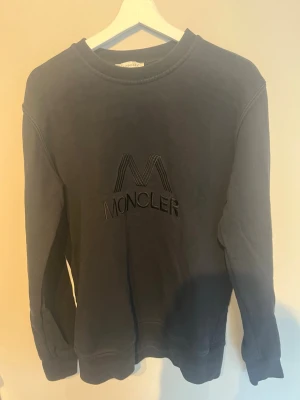 Svart sweatshirt från Moncler - Cool svart sweatshirt från Moncler med stort broderat M och logotyp på bröstet. Tröjan har rund hals, långa ärmar och ribbade muddar. På ryggen finns Moncler-märke vid nacken. Perfekt för dig som gillar exklusiva streetwear-vibes.
