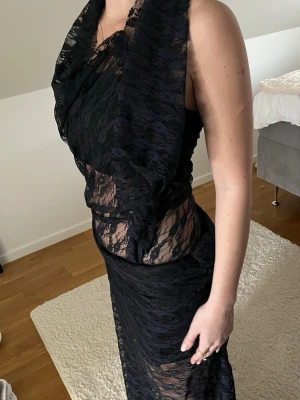 Svart maxi kjol och top  i spets - Elegant svart långklänning med transparent spetsdetaljer och hög krage. Klänningen är ärmlös och har öppen rygg, vilket ger en sofistikerad look. Perfekt för dig som vill sticka ut med en unik och stilren outfit.