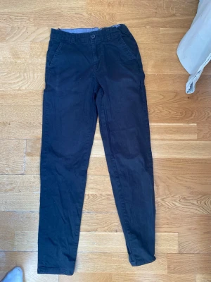 Svarta chinos från Cubus - Säljer ett par svarta chinos med slim fit och klassisk design. Byxorna har snedställda framfickor, bakfickor med knapp och en bekväm midja. Materialet är mjukt och lätt bomull, perfekt för en stilren look. Som man ser på bilderna så är den man spänner med lite sönder 