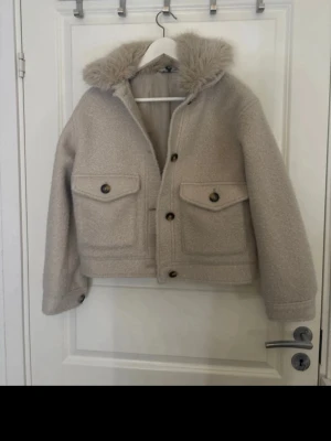 Beige cardiganjacka med fuskpäls - Snygg beige cardiganjacka med fuskpäls på huvan och stora knappar framtill. Jackan har två stora fickor med lock och knapp, samt ribbade muddar vid ärmsluten. Perfekt för kyliga dagar och ger en trendig look.