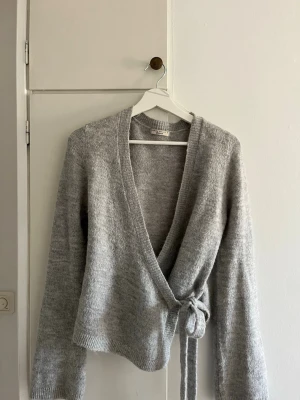 Grå omlottkofta från Gina Tricot - Mysig grå stickad omlottkofta från Gina Tricot med knytband i midjan. Koftan har lång ärm, ribbade muddar och en mjuk, fluffig känsla. Perfekt för lager på lager och enkel att styla till olika outfits.