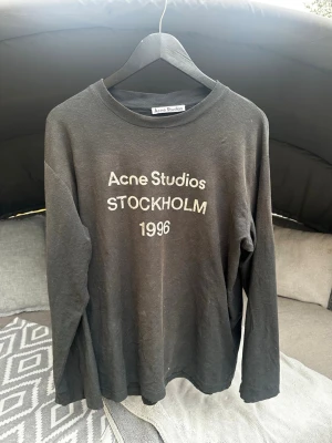 Acne studios t shirt - En stilren och tidlös långärmad tröja från Acne Studios som direkt ger en exklusiv och minimalistisk look. Den klassiska svarta färgen kombineras med det ikoniska trycket Acne Studios Stockholm 1996 framtill – en design som aldrig går ur stil och som passar perfekt i en modern garderob.  Denna tröja är äkta Acne Studios, vilket tydligt syns på originaletiketten och den välgjorda kvaliteten. Perfekt för dig som vill ha ett premiumplagg med skandinavisk designkänsla.