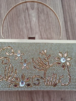 Guldig kuvertväska med pärlor - Snygg kuvertväska i guld med låtsats ädelstenar i blommig design. Väskan har ett metallhandtag och stängs med ett metallspänne. Insidan är fodrad och har ett fack med dragkedja. Väskan har aldrig använts, de fläckar som syns är inte från användning.