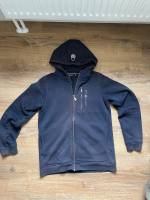 Marinblå hoodie från Sail Racing - Snygg marinblå hoodie från Sail Racing med dragkedja framtill och huva med logga. Tillverkad i mjukt material med borstad insida för extra komfort. Två fickor på sidorna och diskret logotyp på bröstet. Perfekt för en avslappnad och sportig stil.