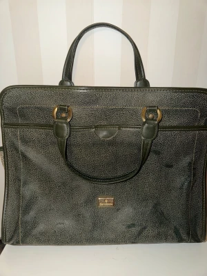 Svart datorväska från Samsonite - Stilren svart datorväska från Samsonite i konstläder med diskret prickigt mönster. Väskan har två rejäla handtag, guldfärgade metalldetaljer och en klassisk rektangulär form. Perfekt för att bära laptop och viktiga papper till plugget eller jobbet.