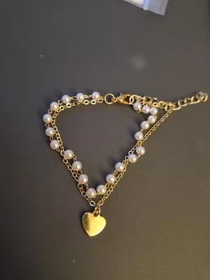 Guldarmband med pärlor och hjärta - Snyggt armband i guldfärgad metall med dubbla kedjor, varav en dekorerad med vita pärlor. Armbandet har ett litet hjärthänge i guldton som detalj. Perfekt accessoar för att lyfta din stil. (Kommer i en box)