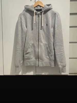 Grå hoodie från Polo Ralph Lauren - Snygg grå zip-hoodie från Polo Ralph Lauren med klassisk blå logga på bröstet. Hoodien har dragsko i huvan, två stora fickor framtill och ribbade muddar. Tillverkad i mjuk bomullsblandning som känns skön mot huden. Perfekt för en avslappnad och stilren look. Finns tecken på slitage men inget större eller som syns för andra när man bär den. Finns de frågor är det bara att fråga 