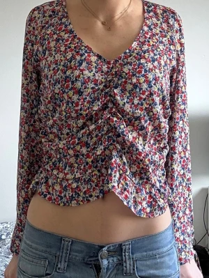 Blommig långärmad blus med rynk - Blus med färgglatt blommönster i rött, blått, vitt och gult. V-ringad hals och rynkdetalj framtill som ger en snygg form. Långärmad modell med tunna band vid ärmsluten. Luftigt material, perfekt till jeans eller kjol. Skriv gärna om ni har frågor eller vill ha fler bilder!!