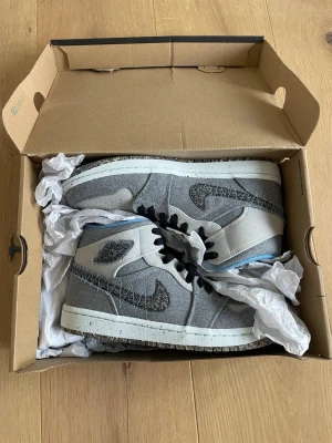 Gråa Nike Air Jordan 1 Mid sneakers - Säljer ett par gråa Nike Air Jordan 1 Mid sneakers med svart swoosh i textil och svarta skosnören. Skorna har blått innerfoder och vit sula med stänkdetaljer. Materialet är en mix av textil och syntet, och modellen har den klassiska Air Jordan-loggan på sidan. Tillverkas inte längre i denna modell. 