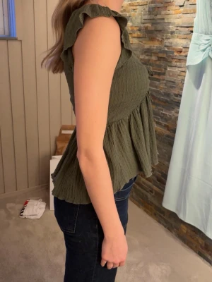 Grön topp med volang och knyt - Hej!🌸Jag säljer en grön topp med volangkantade ärmar och v-ringning. Toppen har ett sött knyt framtill och är tillverkad i ett mjukt, strukturerat bomullstyg. Så snygg till jeans eller kjol. Sparsamt använd, väldigt bra skick!