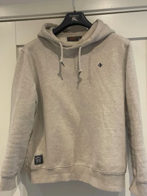 Beige hoodie från Morris  - Snygg beige hoodie från Morris Stockholm med huva och dragsko. Diskret logga broderad på bröstet och patch vid nederkanten. Tillverkad i mjukt bomullsmaterial som känns skönt mot huden. Perfekt för en avslappnad och stilren look.