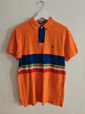 Orange pikétröja från Polo Ralph Lauren - Snygg orange pikétröja från Polo Ralph Lauren med blå krage och knappslå. Tröjan har breda blå, gröna, vita och röda ränder över bröstet samt den klassiska loggan broderad på vänster sida. Perfekt till jeans eller shorts.