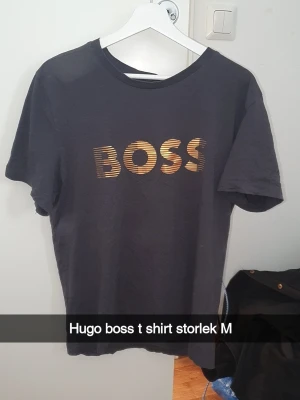 Svart Hugo Boss t-shirt med guldtryck - Snygg svart t-shirt från Hugo Boss med stort guldigt BOSS-tryck på bröstet. Klassisk rund hals och korta ärmar. Perfekt för dig som gillar stilrena plagg med en exklusiv touch.
