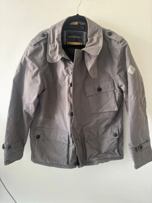 Grå jacka från Henri Loyd. Passar även L - Stilren grå jacka från Henri loyd. med klassisk krage och flera fickor framtill. Jackan har knappar och dold dragkedja samt en patch på ärmen. Perfekt för dig som gillar en enkel men snygg look.