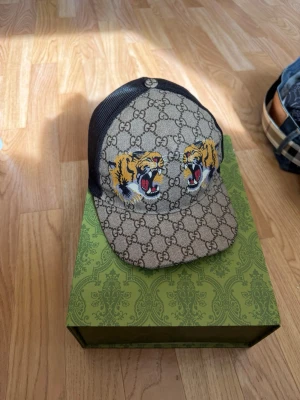 Gucci keps med tigerbrodyr - Säljer en snygg Gucci keps i beige med klassiskt GG-mönster och två broderade tigrar framtill. Kepsen har svart mesh baktill och justerbart band med röd-gröna detaljer. Materialet är canvas och mesh, perfekt för dig som vill sticka ut. Dustbag och box följer med!!! Som ny