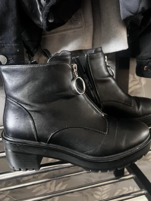 Svarta stövletter med dragkedja - Snygga svarta stövletter i skinnimitation med markerad sula och grov klack. Dekorativ dragkedja med rund metallring framtill ger en cool detalj. Perfekta till jeans eller kjol för en trendig look.