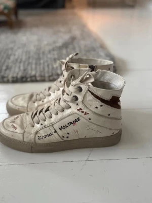 Zadig & Voltaire vita high top sneakers - Unika vita high top sneakers från Zadig & Voltaire med handskrivna detaljer, svarta och röda stjärnor samt text. Skorna har snörning, dragkedja på sidan och bruna detaljer vid hälen. Tillverkade i läder med gummisula och cool streetstil. Bilder är lånade men ser ungefär likadana ut och ändast använt 5 gånger. Ny pris: 3000
