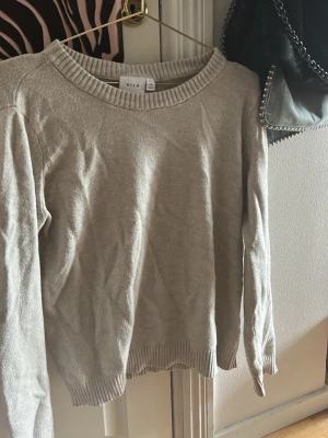 Beige stickad tröja från VILA - En stilren beige stickad tröja från VILA med rund halsringning och ribbade muddar vid ärmslut och nederkant. Perfekt för lager-på-lager och enkel att matcha med jeans eller kjol. Mjuk och skön känsla som passar till många tillfällen.