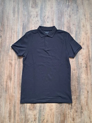 Svart pikétröja från Cotton On - Klassisk svart pikétröja från Cotton On med krage och knappar framtill. Tröjan har korta ärmar och är tillverkad i mjuk bomull, perfekt för en clean och stilren look.