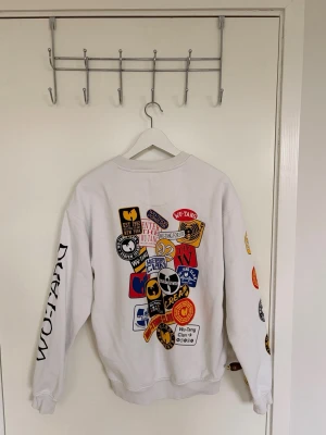 Vit Wu-Tang Clan sweatshirt - Säljer en vit sweatshirt från Wu-Tang Clan med färgglada tryck och patches på ryggen, bröstet och ärmarna. Texten 'WU-TANG FOREVER' syns fram och 'WU-TANG' på ena ärmen. Tröjan har rund hals och ribbade muddar. Perfekt för dig som gillar streetwear och hiphop. Ordinarie pris 700kr.