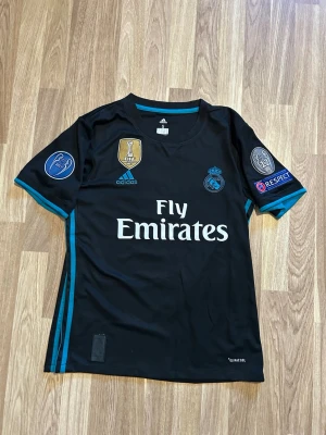 Real Madrid Ronaldo matchtröja svart - Säljer en svart Real Madrid matchtröja från Adidas med Ronaldo 7 på ryggen. Tröjan har turkosa detaljer, Champions League- och FIFA-märken samt Fly Emirates-tryck på bröstet. Perfekt för dig som älskar fotboll och vill ha en ikonisk supportertröja.