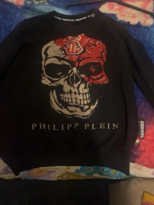 Svart Philipp Plein hoodie med dödskalle - Cool svart hoodie från Philipp Plein med stor dödskalle-tryck i vitt och rött framtill. Tröjan har rund halsringning och ribbade muddar vid ärmslut och nederkant. Perfekt för dig som gillar streetwear och vill sticka ut. Äkta 