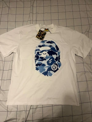 Vit BAPE t-shirt med blått camomönster - Säljer en vit t-shirt från BAPE med det ikoniska apansiktet i blått camomönster på både fram- och baksidan. Klassisk passform och rund hals. Perfekt för dig som gillar streetwear och vill sticka ut med en unik design.