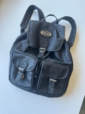 Svart ryggsäck från Daniel Bay - Snygg svart ryggsäck från Daniel Bay i konstläder med två framfickor och justerbara axelband. Väskan har lock med spänne och metallogo framtill. Perfekt för dig som vill ha en stilren och praktisk accessoar.