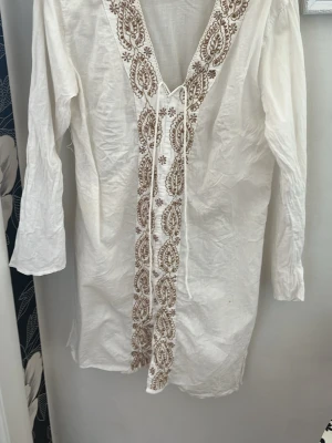 Vit strandtunika med broderi - Säljer en vit strandtunika med långa ärmar och djup V-ringning. Tunikan har vackra beige och bruna broderier längs halsringningen och framtill, samt knytband vid halsen. Tillverkad i ett lätt och luftigt tyg, perfekt att dra över bikinin på stranden.Passar helt klart även en S.
