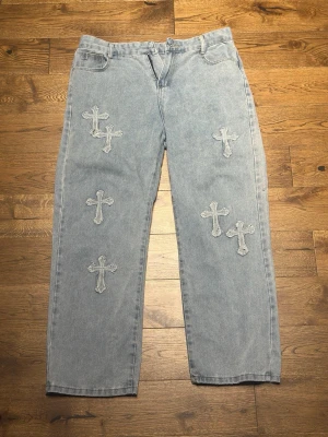 Ljusblå jeans med korsapplikationer - Unika ljusblå jeans med raka ben och flera sydda korsapplikationer både fram och bak. Jeansen har klassisk femficksdesign och en avslappnad passform. Materialet är denim i bomull och detaljerna ger ett edgy och trendigt uttryck.