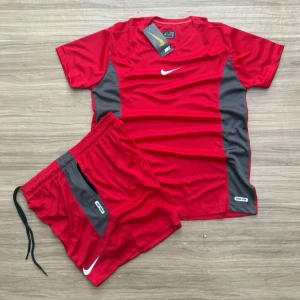 Rött Nike träningsset herr - Snyggt rött träningsset från Nike med t-shirt och shorts. Setet har gråa paneler på sidorna och vita Nike-loggor på både tröjan och shortsen. Perfekt för gymmet eller löpning. Materialet är lätt och andas bra.