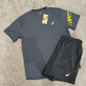  Asics sett  - Snyggt träningsset från Asics med en grå t-shirt med vit logga och gul detalj på ärmen samt svarta shorts med vit logga. Perfekt för gym eller löpning, tillverkat i funktionsmaterial som andas.