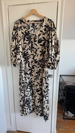 Beige långklänning med svart blommönster - Säljer en beige långklänning från Lindex med svart blommigt mönster. Klänningen har v-ringning, puffiga trekvartsärmar och en avslappnad passform. Perfekt för dig som gillar statement-plagg med snygga detaljer.
